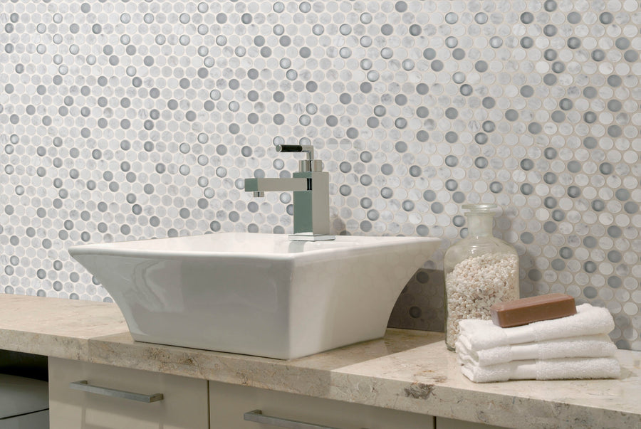 The Luna Penny Round features a modern bathroom sink with a square white basin, chrome faucet, stacked white towels, bar soap, and a glass jar of cotton balls on a stone countertop, set against a classic penny round mosaic tile wall.