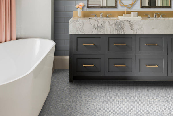 Modern bathroom featuring a white freestanding tub, Luna Penny Round tiles on the floor, gray double-sink vanity with gold handles, marble countertop, gray paneled wall, and a pink curtain for a classic touch.