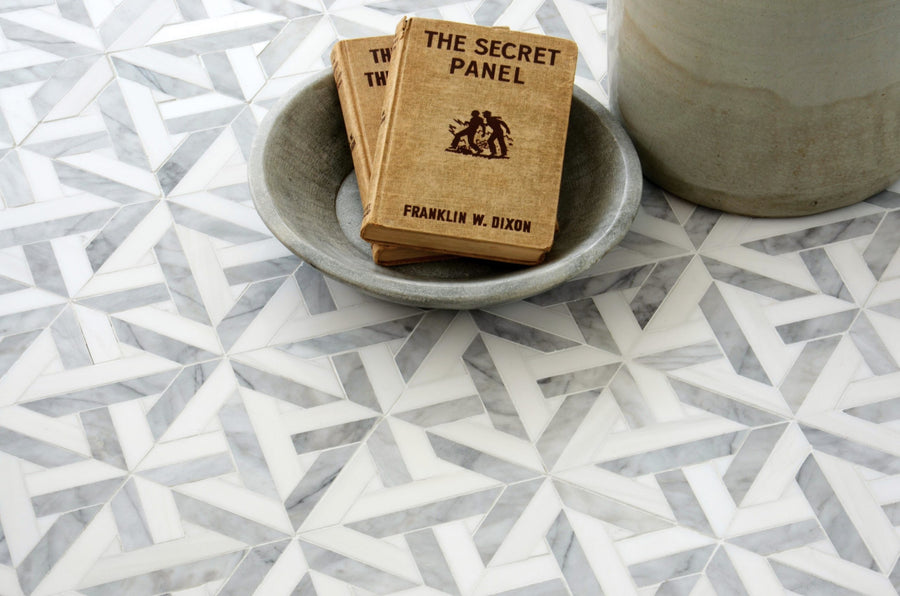 A small stack of vintage books in a gray bowl sits on a Talya Marmara 9 11/16 x 9 11/16 Waterjet Natural Stone tiled surface, next to a large ceramic vessel.