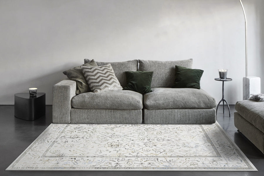 A modern living room features a gray sofa with green and patterned pillows, the Merit Area Rug in light geometric style, a small black side table with a cup, a candle on a black stand, and a white curved floor lamp against a gray wall.