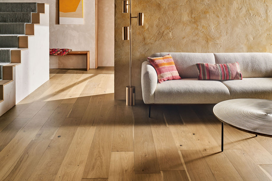 A modern living room with Monticello 8 1/2" White Oak Hardwood floors featuring natural knots, a beige curved sofa with colorful pillows, a round coffee table, gold lighting, and a textured tan wall by stairs and a hallway bench with red blankets.