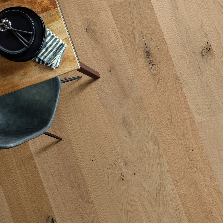 A top-down view of a wooden table with natural knots, a plate, fork, and striped napkin beside a dark chair. The table is placed on Monticello 8 1/2" White Oak Hardwood flooring with visible grain and character.