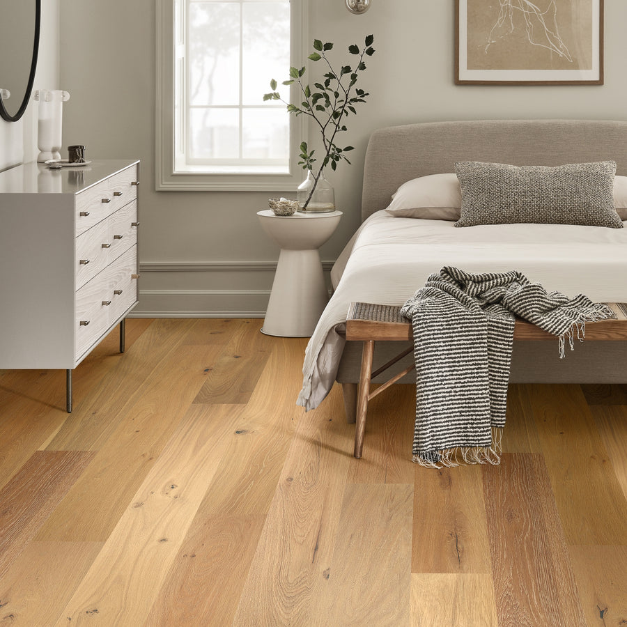 A modern bedroom with Monticello 8 1/2" White Oak Hardwood flooring, a beige upholstered bed, white bedding, gray throw, wood bench, white dresser, round mirror, potted plant, and a sunlit window.