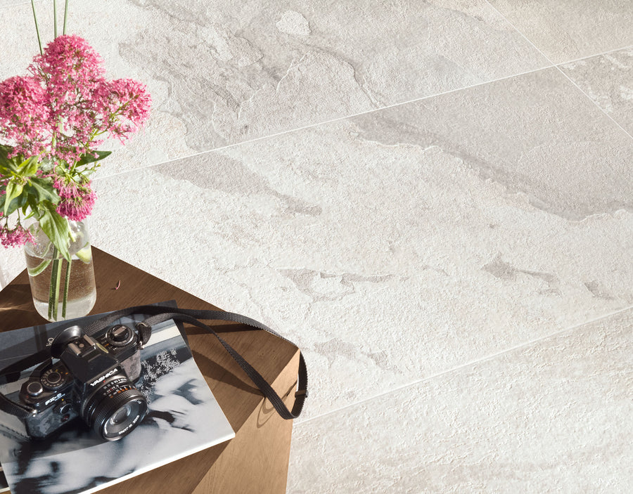 A close-up of a light gray More 12x24 Porcelain Tile floor with a slate look. A wooden table in the foreground holds pink flowers in a vase, a vintage camera, and a black-and-white photo.