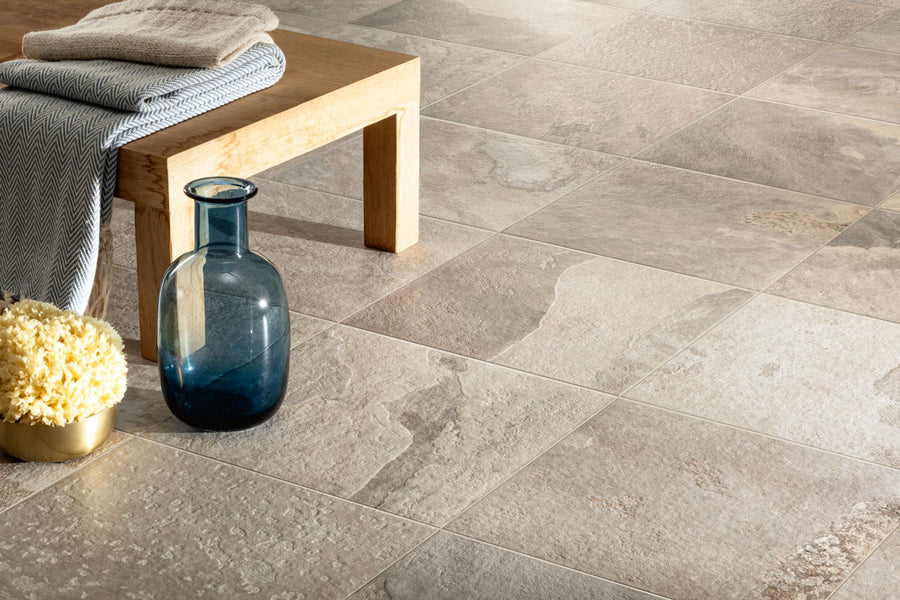 A close-up of More 12x12 Porcelain Tile in beige used as outdoor flooring, with a light wooden bench, folded cloth, blue glass vase, and a yellow sponge in a gold bowl arranged in the corner.