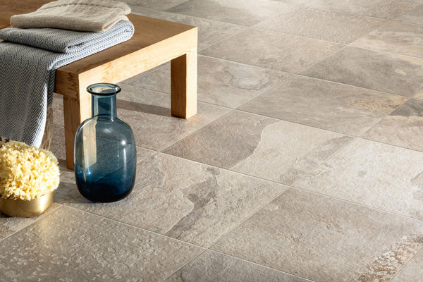 A close-up of More 12x12 Porcelain Tile in beige used as outdoor flooring, with a light wooden bench, folded cloth, blue glass vase, and a yellow sponge in a gold bowl arranged in the corner.