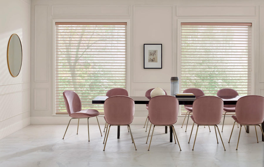 A modern dining room features a black table, eight pink velvet chairs with gold legs, large windows with Nantucket™ Window Shadings for soft light, a round mirror, and a framed picture on light walls.