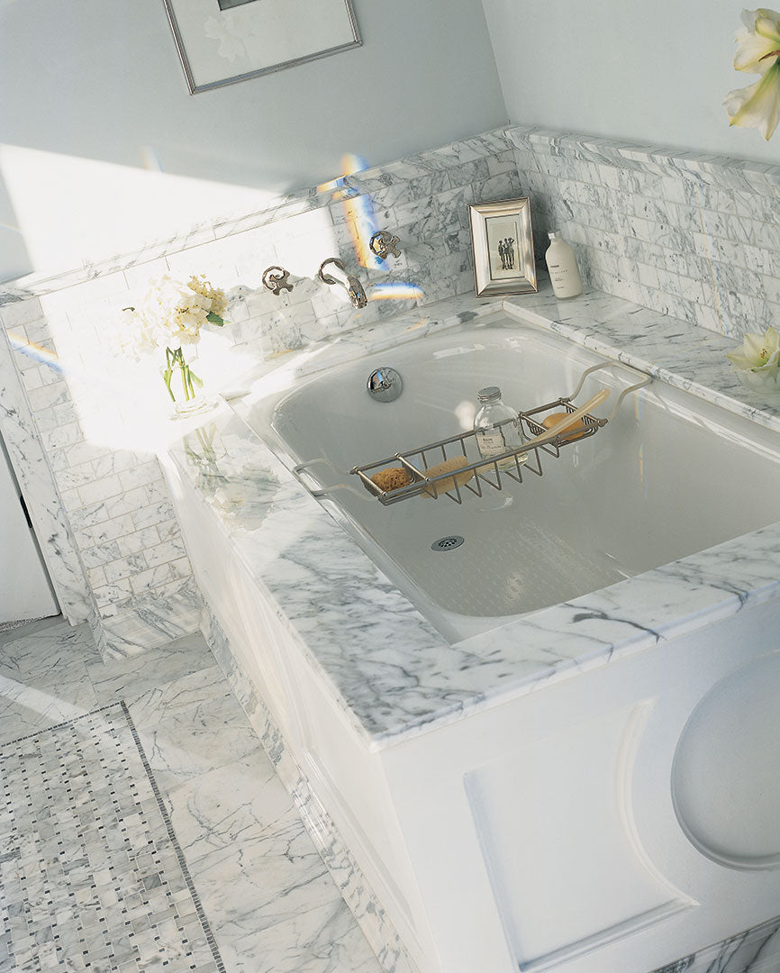 A luxurious bathroom showcases a built-in tub with a gold tray, surrounded by White Carrara Select 3x6 Marble mosaics, decorative accents, flowers, and framed pictures on the ledge.