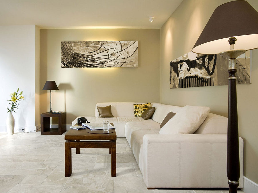 A modern living room features a white sectional sofa, wooden coffee table, floor lamp, and abstract art on beige walls. A vase with flowers rests by the window, while elegant Diana Royal 18x18 Antique Marble floors anchor the space.