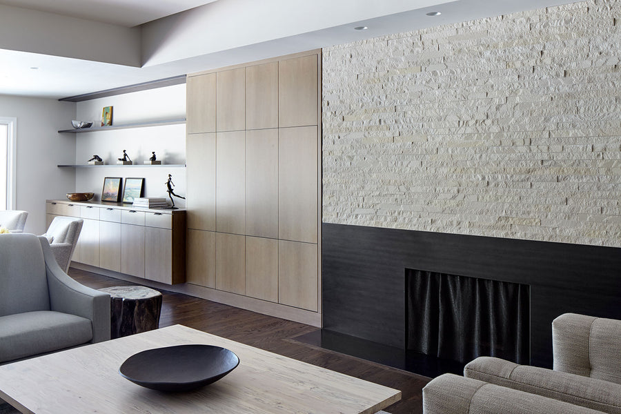 A modern living room features light wood cabinets, open shelves with decor, a Strip Cladding 2x12 Natural Stone Ledger Panel accent wall and dark fireplace, neutral armchairs, and a light wood coffee table topped with a black bowl.