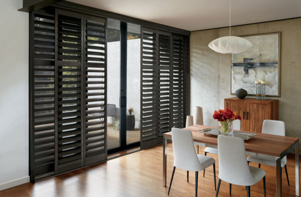 A modern dining room with a wooden table, four white chairs, red flowers in a vase, abstract wall art, and large NewStyle® Hybrid Shutters on glass doors. The space features light wood floors and a white pendant lamp.
