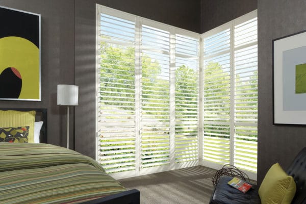A modern bedroom with large corner windows fitted with affordable NewStyle® Hybrid Shutters that allow natural light to filter in. The space features a striped bedspread, contemporary art, neutral walls, and a view of trees outside.