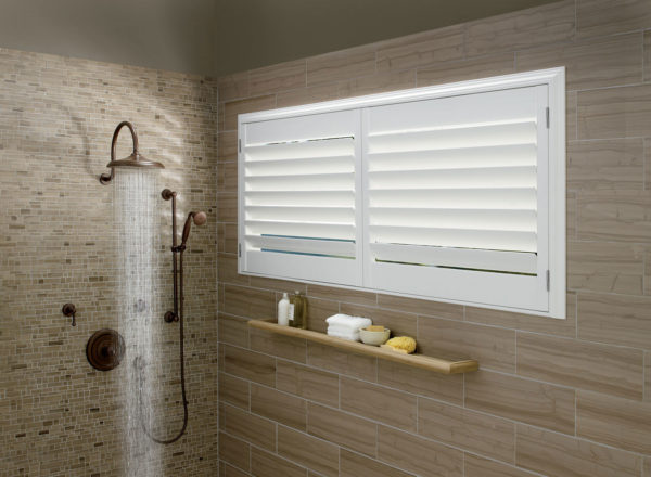Modern bathroom with a walk-in shower featuring a rainfall showerhead and beige tile walls. A shelf holds toiletries below a frosted window fitted with stylish, affordable NewStyle® Hybrid Shutters in classic white.