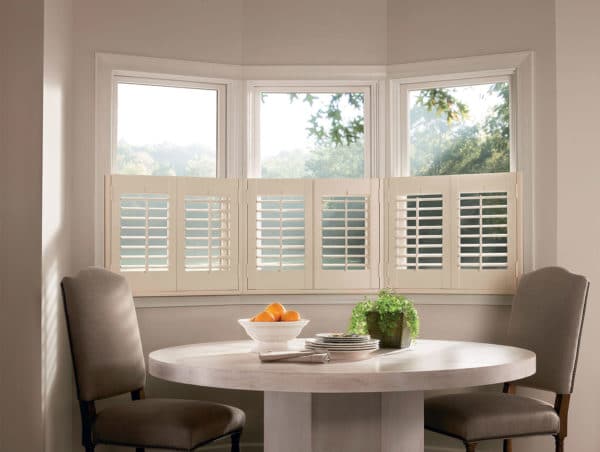 A round dining table with two chairs sits by a bay window featuring affordable NewStyle® Hybrid Shutters. On the table are a bowl of oranges, a potted plant, and plates, while trees can be seen outside the window.