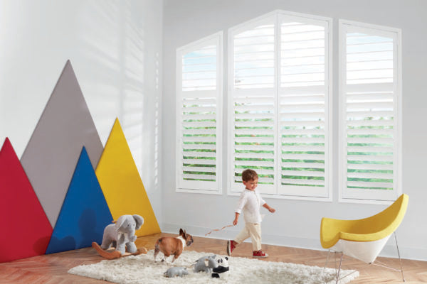 A young child walks in a bright, modern room featuring large NewStyle® Hybrid Shutters, playful mountain-shaped wall decor, stuffed animals, a dog, and a yellow chair on a fluffy rug.