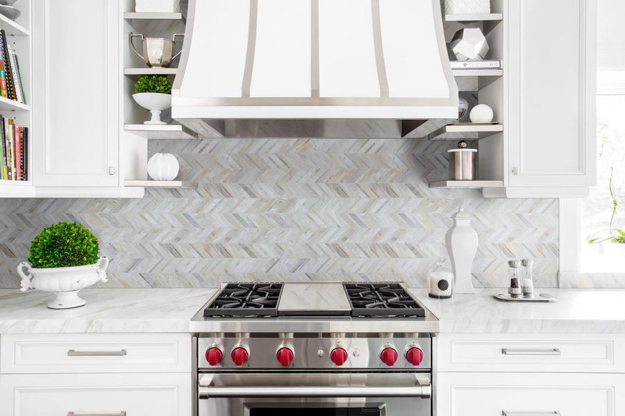 Modern kitchen with white cabinets, natural marble countertops, a stainless steel stove with red knobs, a white range hood, and an Olympia Sandalwood Chevron Marble Mosaic backsplash. Decorative items and plants adorn the shelves.