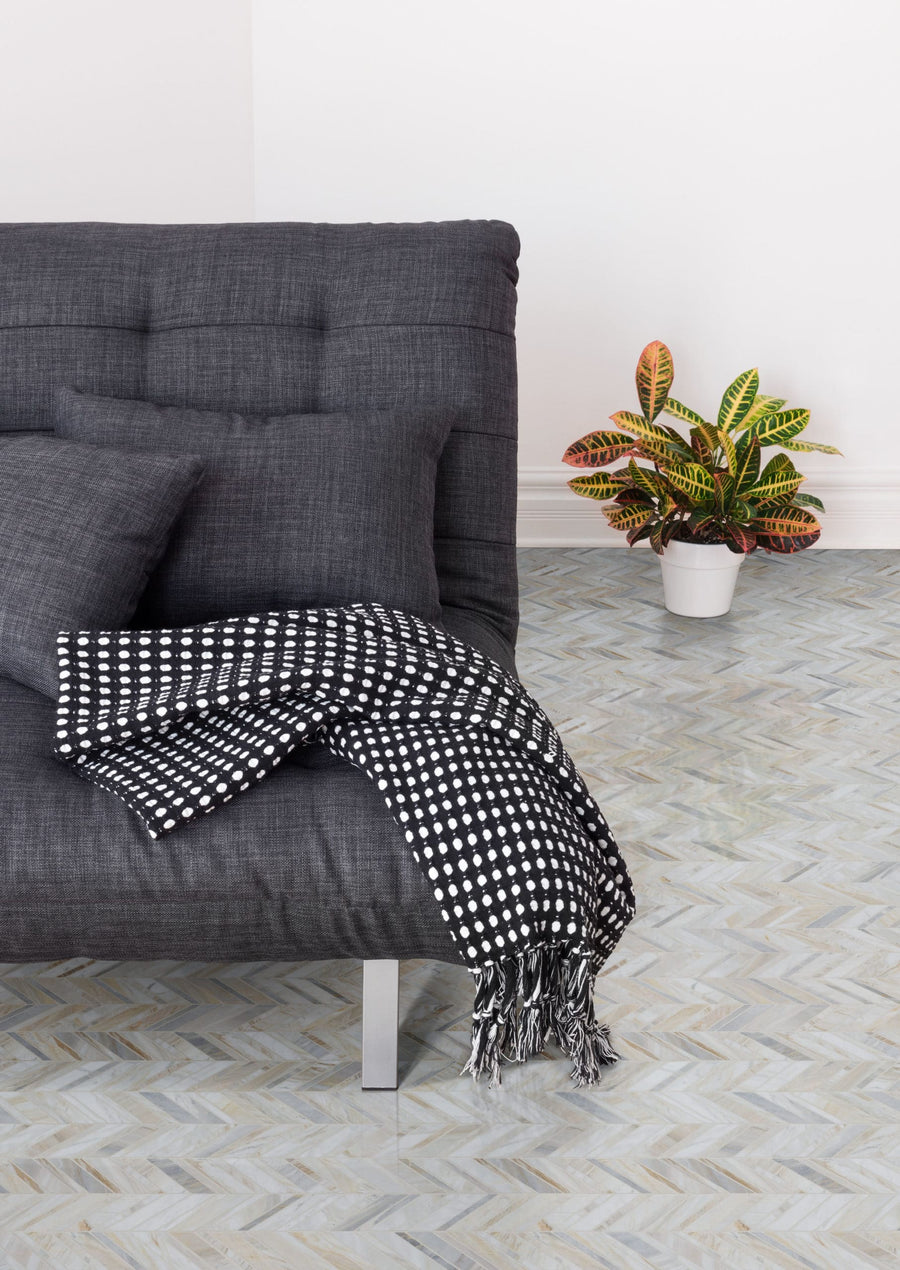 A modern gray sofa with two pillows, a black-and-white polka dot throw, and a small potted plant sits on Olympia Sandalwood Chevron Marble Mosaic tiles.