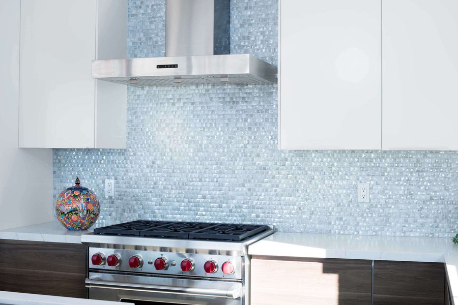 A modern kitchen with a stainless steel stove with red knobs, matching range hood, glossy white cabinets, an Origami 1x1 Glass Mosaic Tile blue backsplash, and a colorful mosaic vase on the counter.