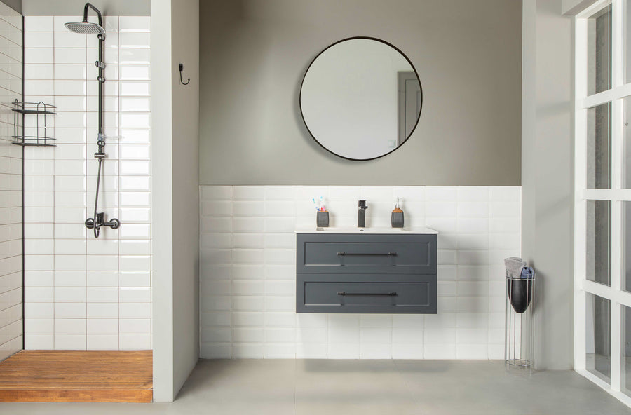 Modern bathroom with a walk-in shower on the left and a Nova 36" Vanity in dark gray, wall-mounted with two drawers and a round mirror above. Gray and white tiled walls complement the modern countertop displaying toiletries.