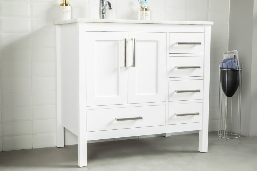 The Ashley 36" Vanity in natural oak has metal handles, two center doors, five side drawers, and a lower drawer. It's set against white tiled walls with gray flooring and is accompanied by a metal basket filled with towels.
