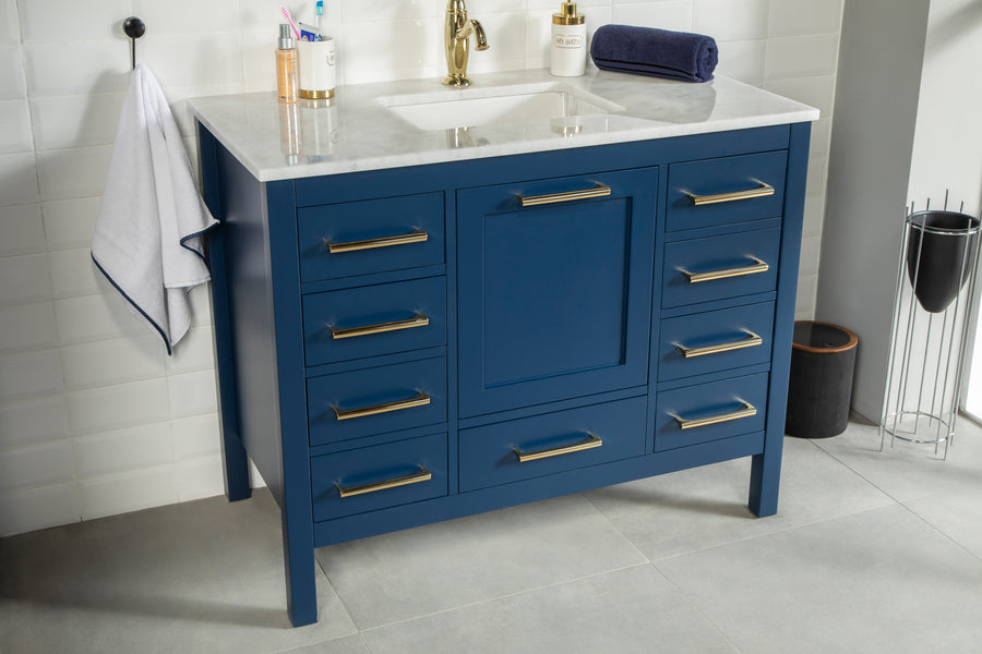 The Ashley 42" Vanity features a blue cabinet with gold handles, a white marble countertop with built-in sink, and is styled with a white towel on the side, toiletries on the counter, and white subway tiles on the wall.