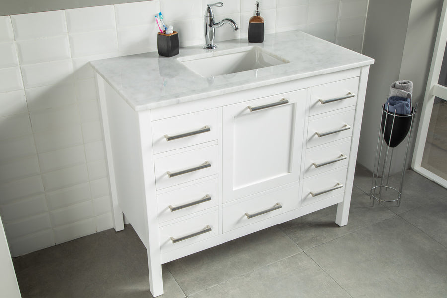 The Ashley 42" Vanity features a white finish, marble countertop, built-in sink, chrome faucet, and spacious drawers. It stands on a gray tiled floor next to a black towel holder.