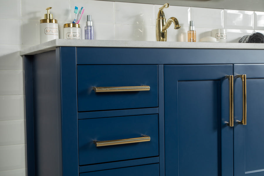 The Ashley 48" Vanity features a blue finish with gold handles, topped with a white countertop and gold faucet. Toiletries such as toothbrushes, toothpaste, soap dispensers, and skincare items sit on the counter against a white tiled wall.