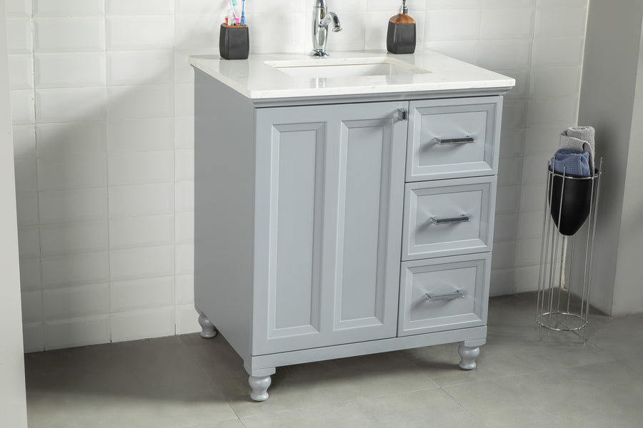 The Isabel 30" Vanity features solid wood drawers, a cabinet, and short legs, topped with a white sink and faucet. It’s complemented by toothbrush holders, soap, and a towel holder beside it against the white tiled wall.