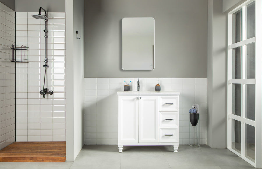 Modern bathroom with a glass shower on the left, white tiled walls, and an Isabel 36" Vanity with solid wood drawers and a mirror above. A trash bin is next to the sink, while natural light enters through large right-side windows.