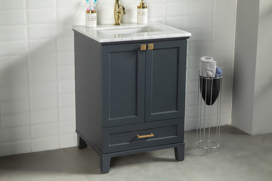 The Paloma 24" Vanity features a dark blue cabinet, gold hardware and faucet, white countertop, and soft close drawers. A small metal towel rack with rolled towels stands beside it against a white tiled wall.