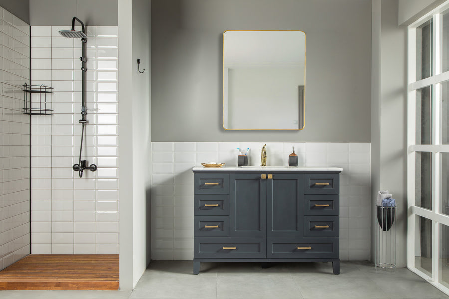 Modern bathroom with a walk-in shower on the left, white subway tiles, and wood floors. Centered is a Paloma 48" Vanity with soft-close drawers and gold handles beneath a square mirror. A trash can sits to the right.