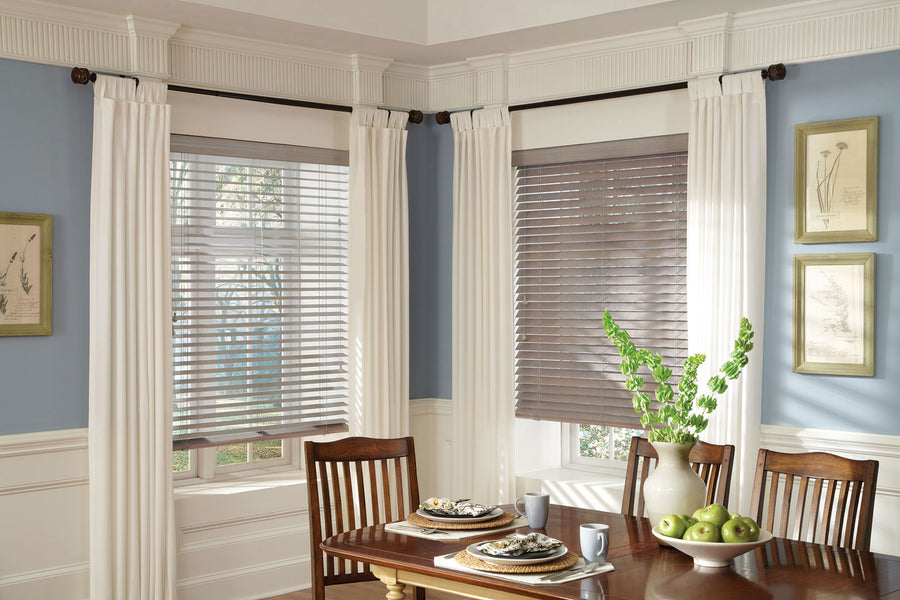 A dining area with a wooden table set for two and matching chairs. The windows feature Parkland® Wood Blinds with white curtains. Green apples and a vase with leaves decorate the table, while botanical prints hang on the walls.