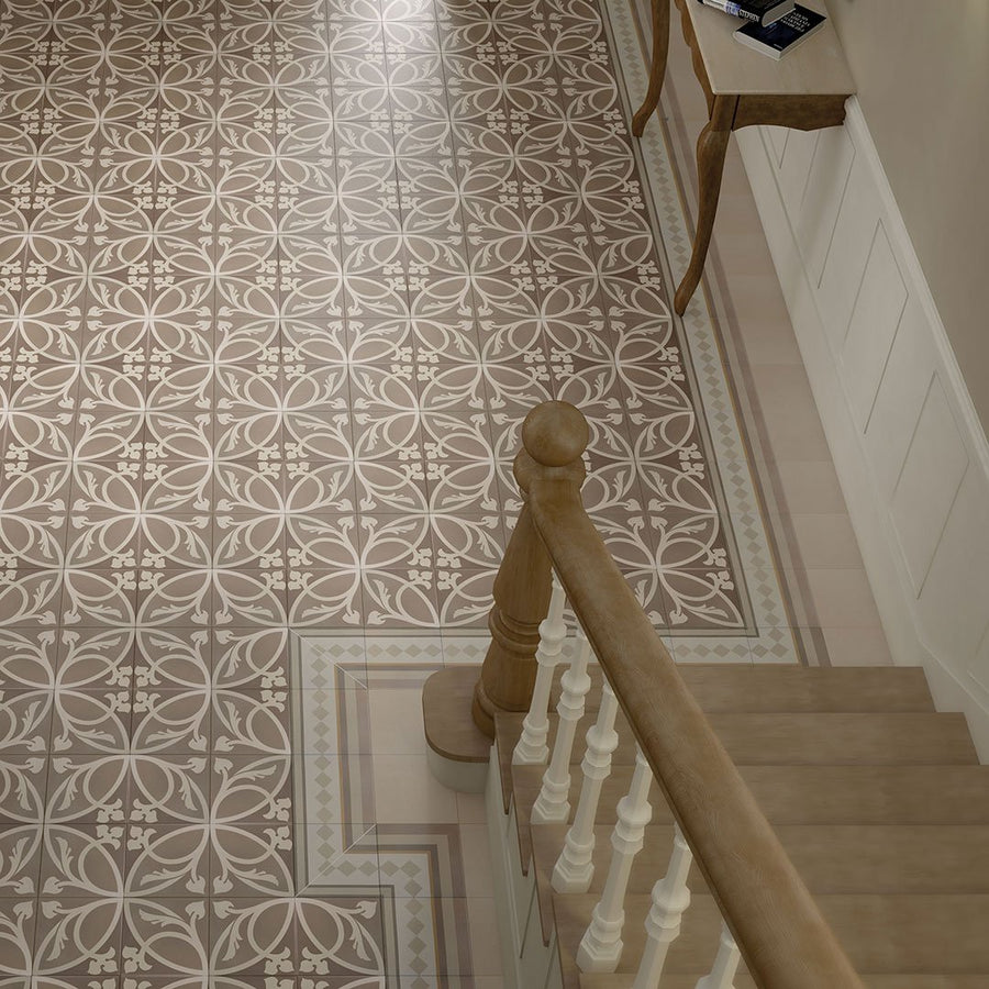 A hallway with Caprice Liberty Taupe Patterned Tile in beige and brown, featuring floral and geometric designs. A wooden staircase with white spindles and a small wooden console table sit along the wall.