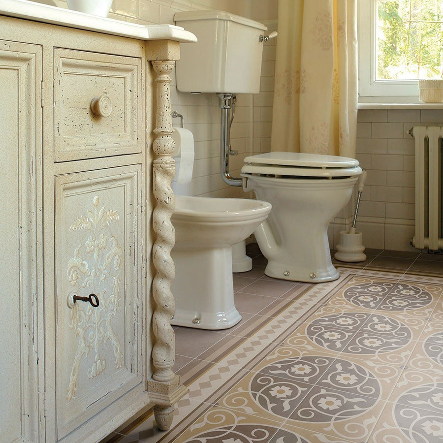 A vintage bathroom features an ornate cream vanity, toilet, bidet, and Caprice Loire Patterned Tile flooring beneath a sunlit window.