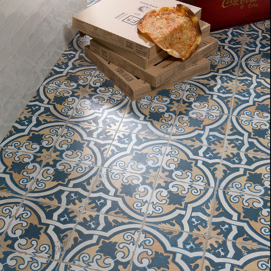 A slice of pizza rests on stacked boxes atop Kings Canarsie 17 3/4 x17 3/4 Patterned Tile, featuring blue, beige, and white designs near a white tile wall.