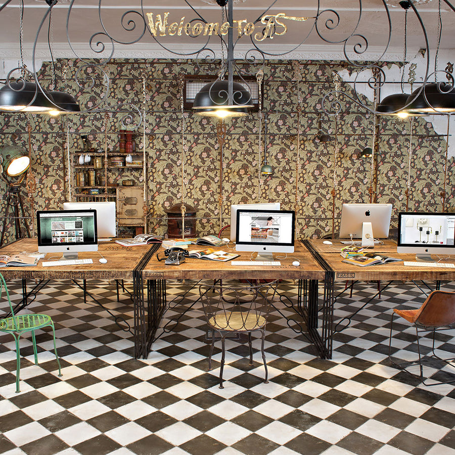 Open workspace with wooden desks, computers, and mismatched chairs on Kings Damero 17 3/4 x17 3/4 patterned tile flooring. Floral wallpaper, vintage decor, and a Welcome sign evoke a cozy eclectic vibe with touches of vintage industrial design.