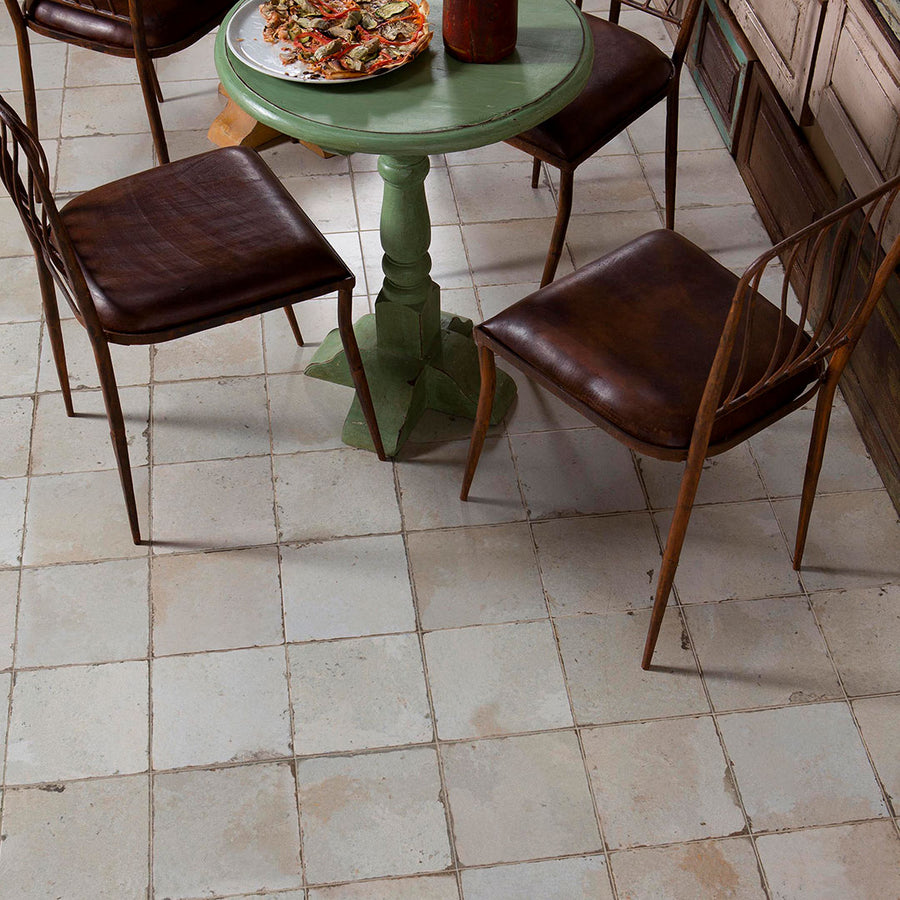 A small green round table with a plate of food and three brown leather chairs sits on Kings Manhattan 17 3/4 x17 3/4 Patterned Tile, giving the space a vintage industrial vibe with its worn, rustic appearance.