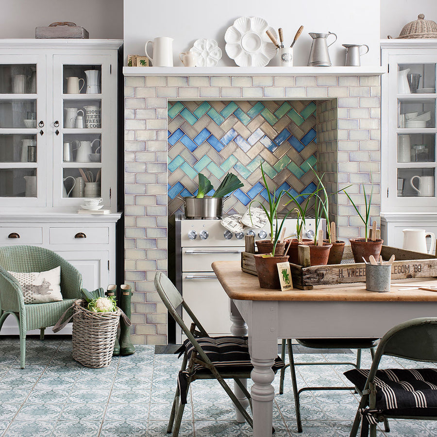 A cozy farmhouse kitchen with white cabinets, vintage dishes, and ceramic floor and wall tile in blue and white patterns like the Kings Seagate 17 3/4 x 17 3/4 Patterned Tile, plus green chairs and a stove set in a tiled herringbone alcove.