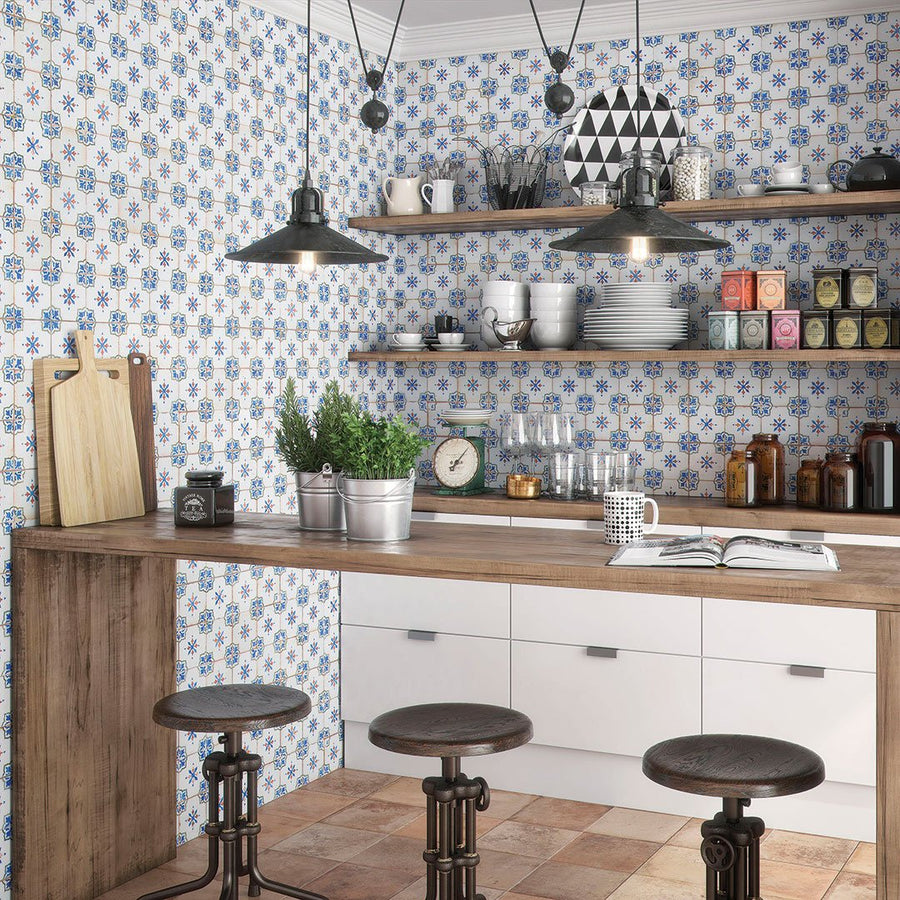 A cozy kitchen features Mirambel 13x13 Patterned Tile on the walls, a wooden counter with three stools, open shelves with dishes and jars, hanging lights, plus herbs, a mug, and cutting boards on the counter.