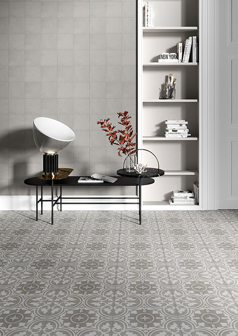 A modern living room features Reverie #11 8x8 patterned tile flooring, a black coffee table with a decorative lamp and red-leaf vase, a built-in white bookshelf with books and decor, and a gray tiled accent wall.