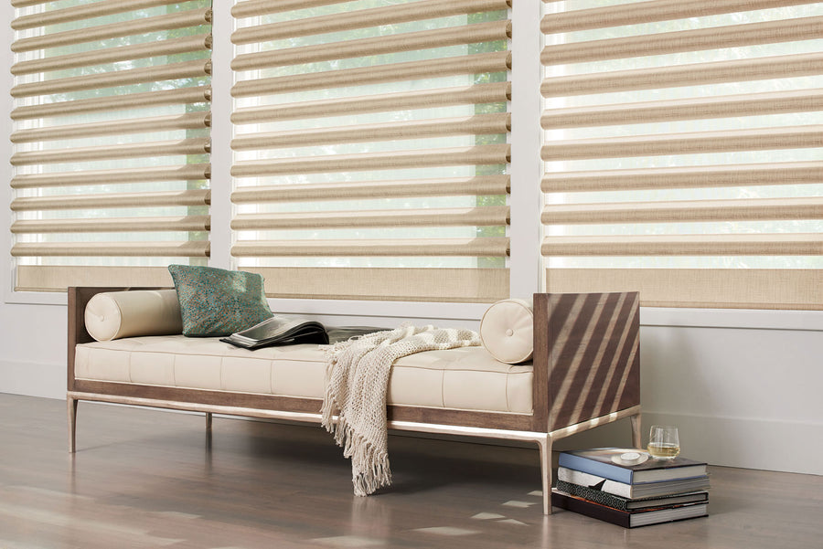 A modern cream and wood daybed with pillows, a knit throw, and an open book sits by large windows fitted with Pirouette® Window Shadings for light filtration and UV protection; stacked books and a glass of water rest beside it.