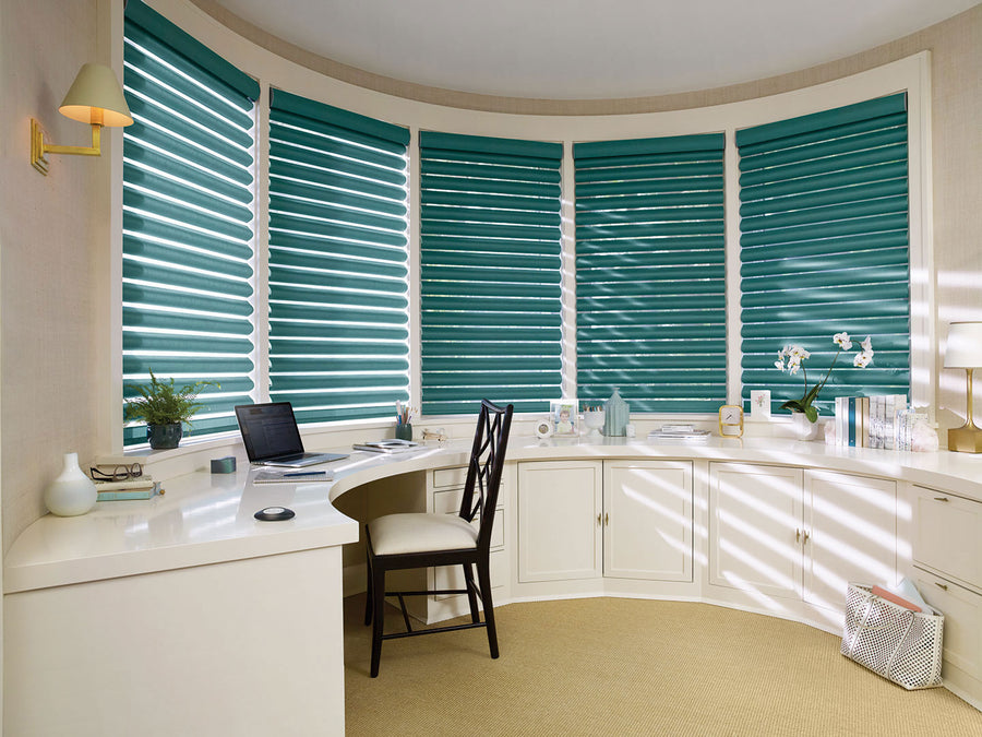 A bright, curved home office features teal Pirouette® Window Shadings with PowerView® Motorization for perfect light control on large windows, a white built-in desk, chair, laptop, books, plants, and decor with sunlight streaming in.