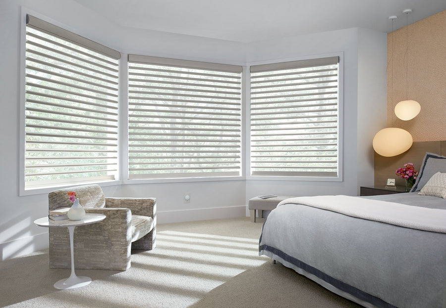 A bright, modern bedroom with large windows features Pirouette® Window Shadings with PowerView® Motorization for versatile light control, a bed with gray and white bedding, a round side table with a vase, an armchair, and two globe pendant lights.