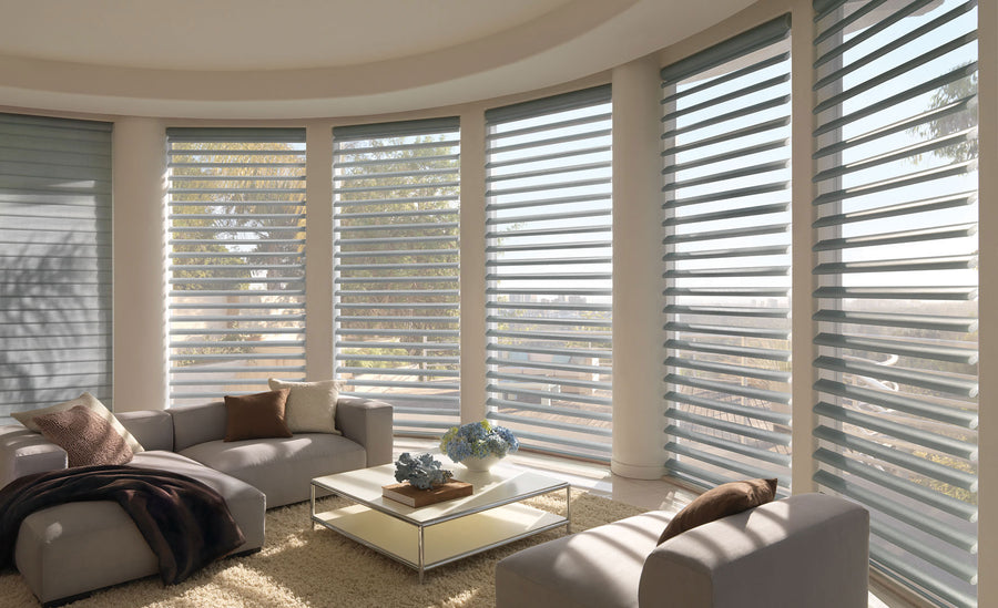 A modern living room with large curved windows features Pirouette® Window Shadings with PowerView® Motorization, beige sofas, a glass coffee table topped with flowers and decor, and softly filtered light for a bright, airy feel.