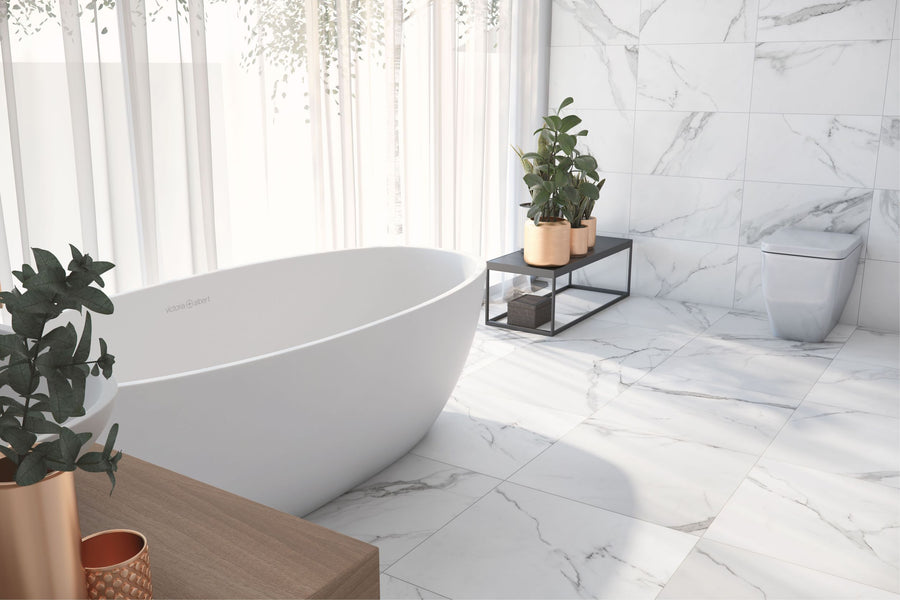 A modern bathroom features Buckingham Palace 12x24 Porcelain Tile on floors and walls, a freestanding oval bathtub, white toilet, green potted plants, and large sheer curtains allowing in natural light.