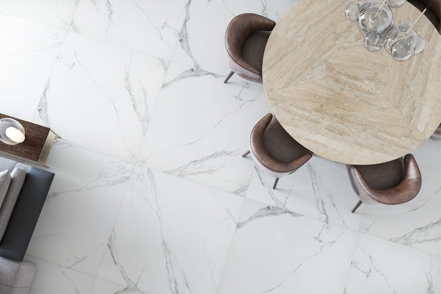 Buckingham Palace 24x24 Porcelain Tile flooring with gray veining, a round light wood dining table, four brown cushioned chairs, a partial gray sofa, and a decorative bowl on a nearby side table.