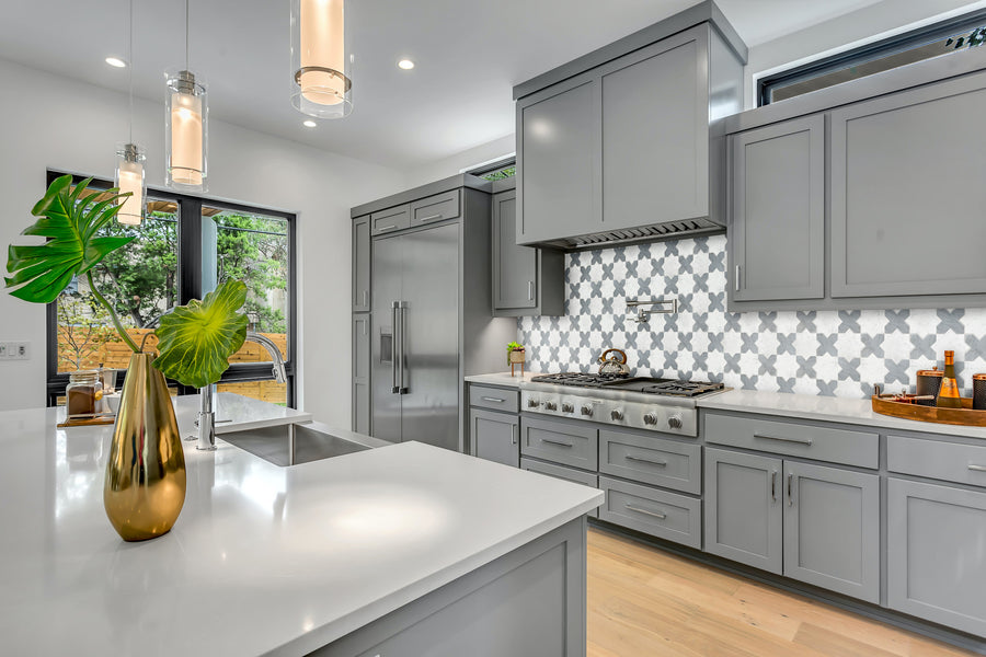 Modern kitchen with gray cabinets, stainless steel appliances, Casablanca Marble & Stone Mosaic backsplash, white Carrara marble countertops, pendant lights, and a gold vase with green leaves on the island. Large windows offer natural light and outdoor views.