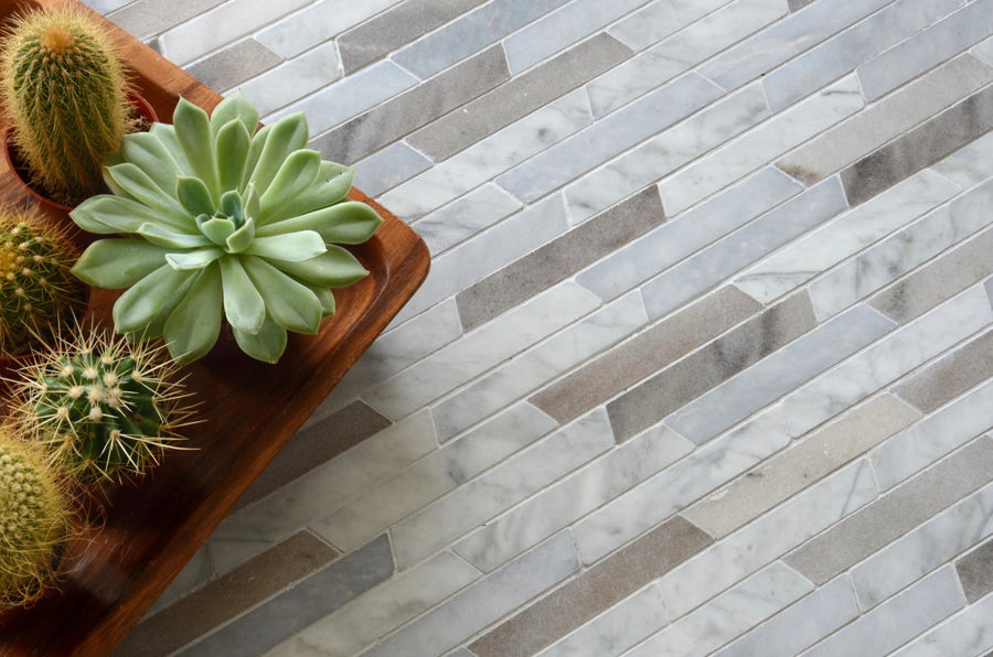 A wooden tray with three small cacti and a succulent sits on Talya Rhodes 8 13/16 x 8 13/16 Waterjet Natural Stone tile in a light gray and white herringbone pattern, inspired by modern marble and stone geometries. Plants are arranged to the left.