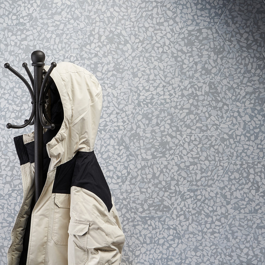 A cream-colored hooded jacket with black accents hangs on a black coat rack against a light blue patterned wall and Riazza 9" Hexagon Porcelain Tile.