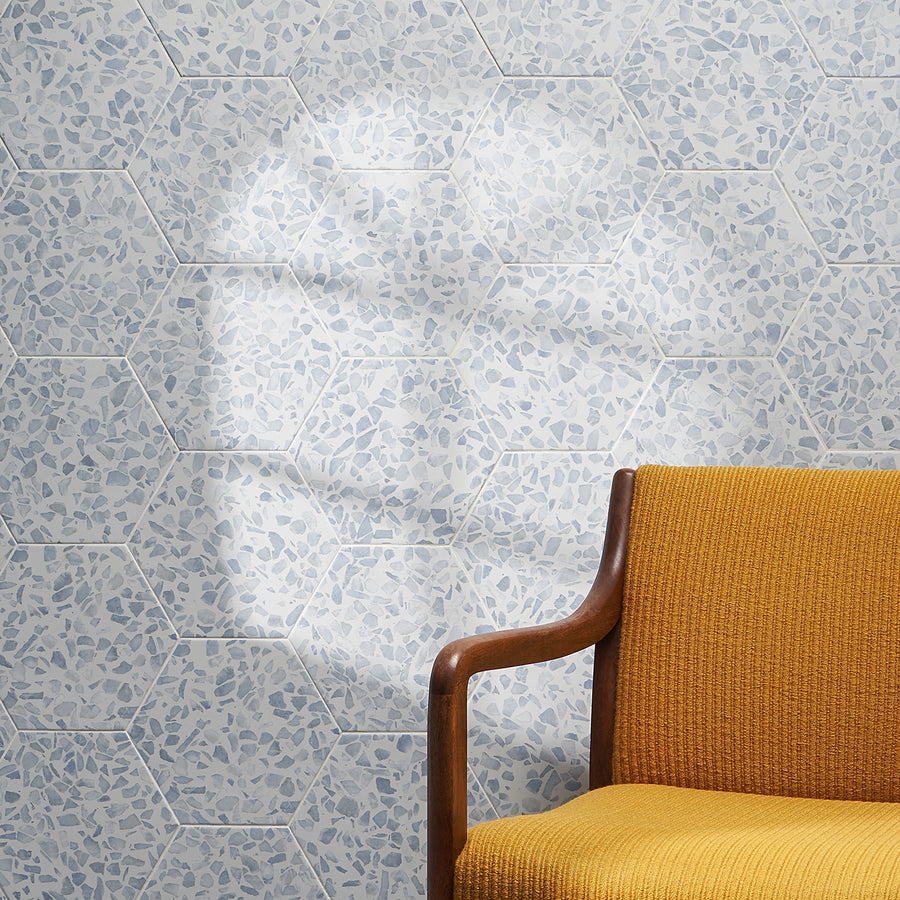 A mustard yellow upholstered chair with wooden armrests sits before a wall of Riazza 9" Hexagon Porcelain Tile in blue and white. Sunlight casts a circular glare, accentuating the matte finish and unique terrazzo pattern.